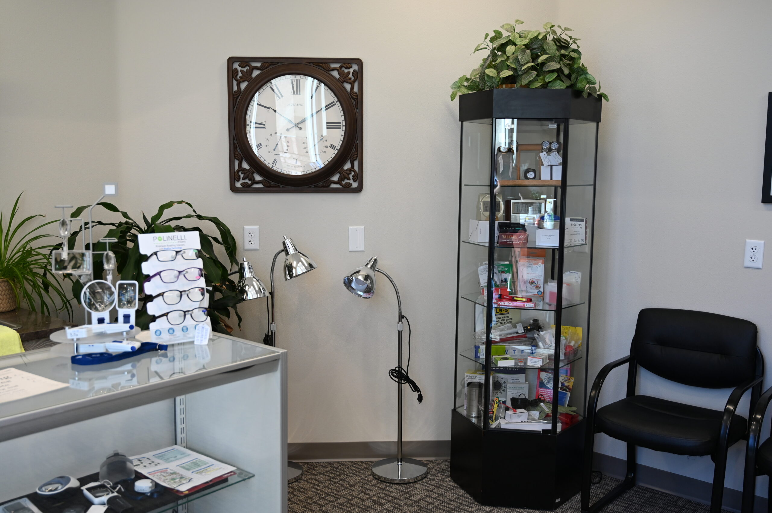 The image shows the interior of a visually rehabilitative skills center. There is a display cabinet filled with various products related to vision aids, such as magnifiers and eyeglasses. To the left of the cabinet is a black chair and a set of three chrome floor lamps. A large decorative clock with Roman numerals is mounted on the wall above the lamps.