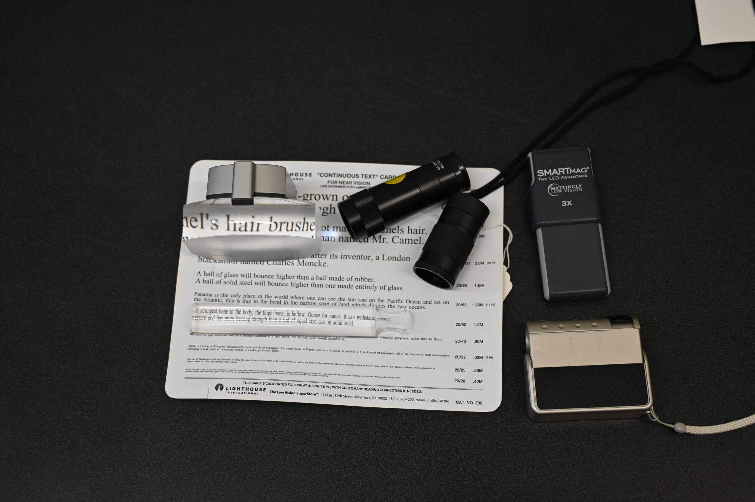 The image shows an array of visual aids and magnification devices placed on a dark surface. There is a reading card with various text sizes for near vision. Several magnifiers and vision tools are arranged on and around the card, including a handheld magnifier, and a couple of other compact magnification devices. These tools are likely used to assist individuals with low vision in reading and other detailed tasks.