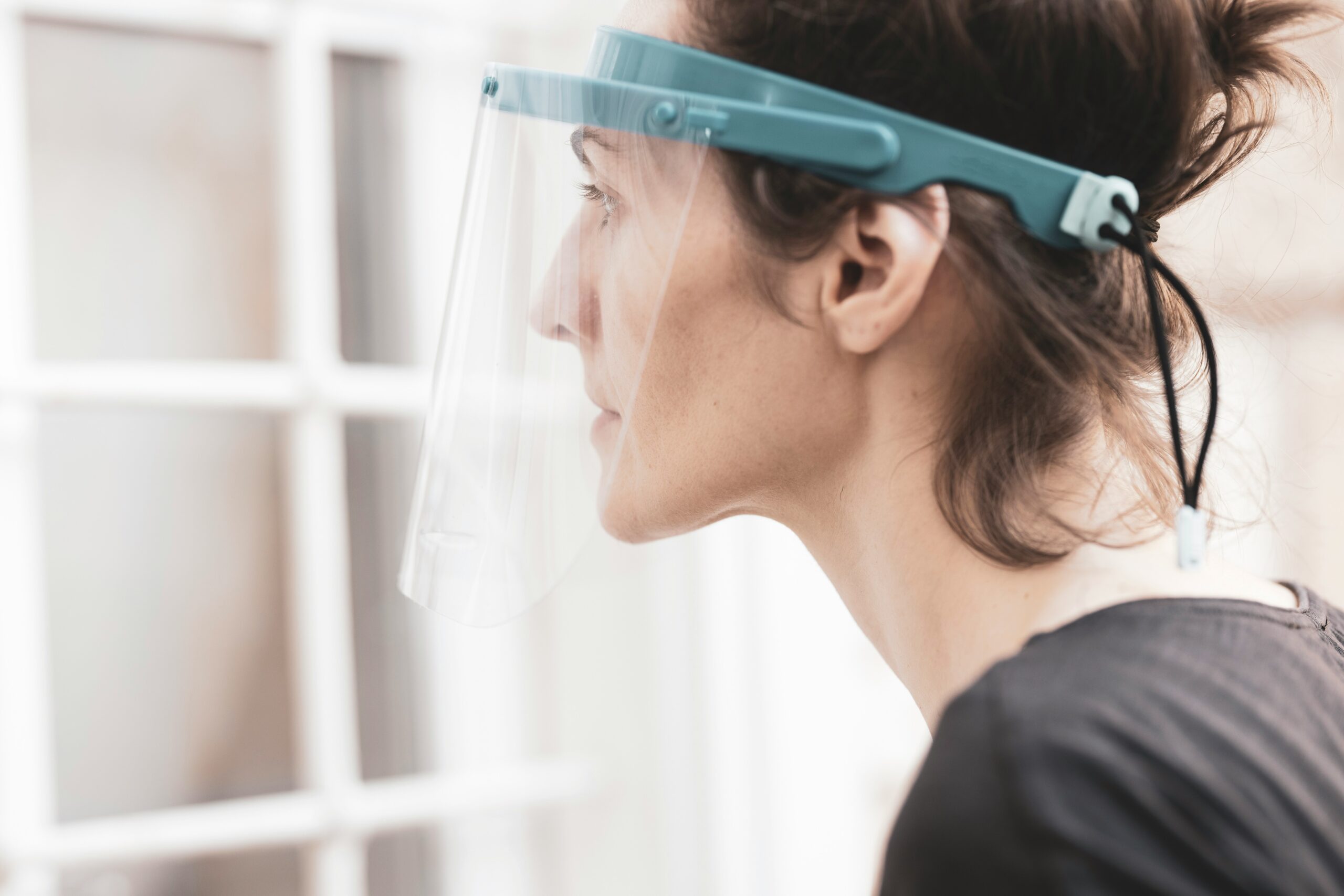 The image shows a side profile of a person wearing a transparent face shield with a light blue headband. The shield covers their face, providing protection, and the person has their hair tied back loosely.