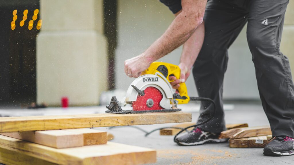 The image shows a person using a yellow Dewalt circular saw to cut through a piece of wood. The person is bent over, holding the saw with both hands, with wood dust flying around as the saw cuts.