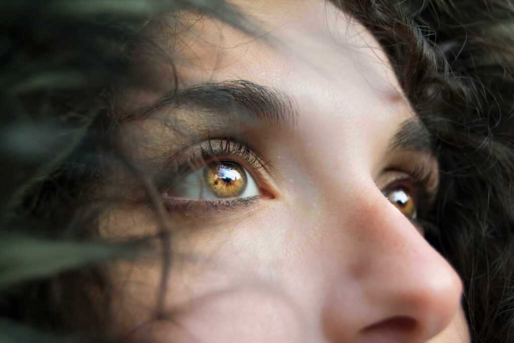 The image is a close-up of a person’s face, focusing on their eyes and upper part of their nose. The person's golden-brown eyes are looking upwards, with light reflecting in their pupils, possibly showing a reflection of the sky or trees