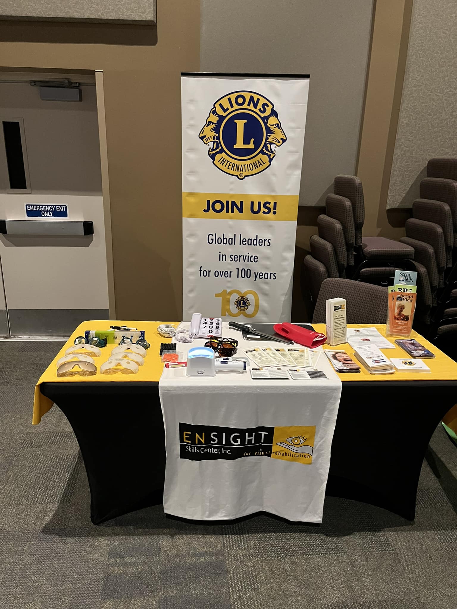 The image depicts an informational booth inside a room. On the left, there is a vertical banner with the Lions International logo and the words "JOIN US! Global leaders in service for over 100 years." A table is covered with a yellow cloth on the left side and a black cloth on the right side, and it bears the logo "ENSIGHT Skills Center, Inc. for visual rehabilitation."