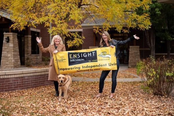 A woman  is holding a banner with her guide dog. There is another woman holding the right side of the banner. They are standing in a courtyard surrounded by leaves.