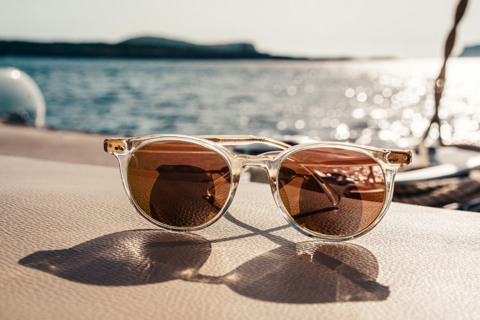 A pair of sunglasses with clear, translucent frames and brown-tinted lenses resting on a wooden boat deck. The sunglasses cast a distinct shadow on the textured wood surface. In the background, a body of water stretches toward distant hills or mountains under an overcast sky.