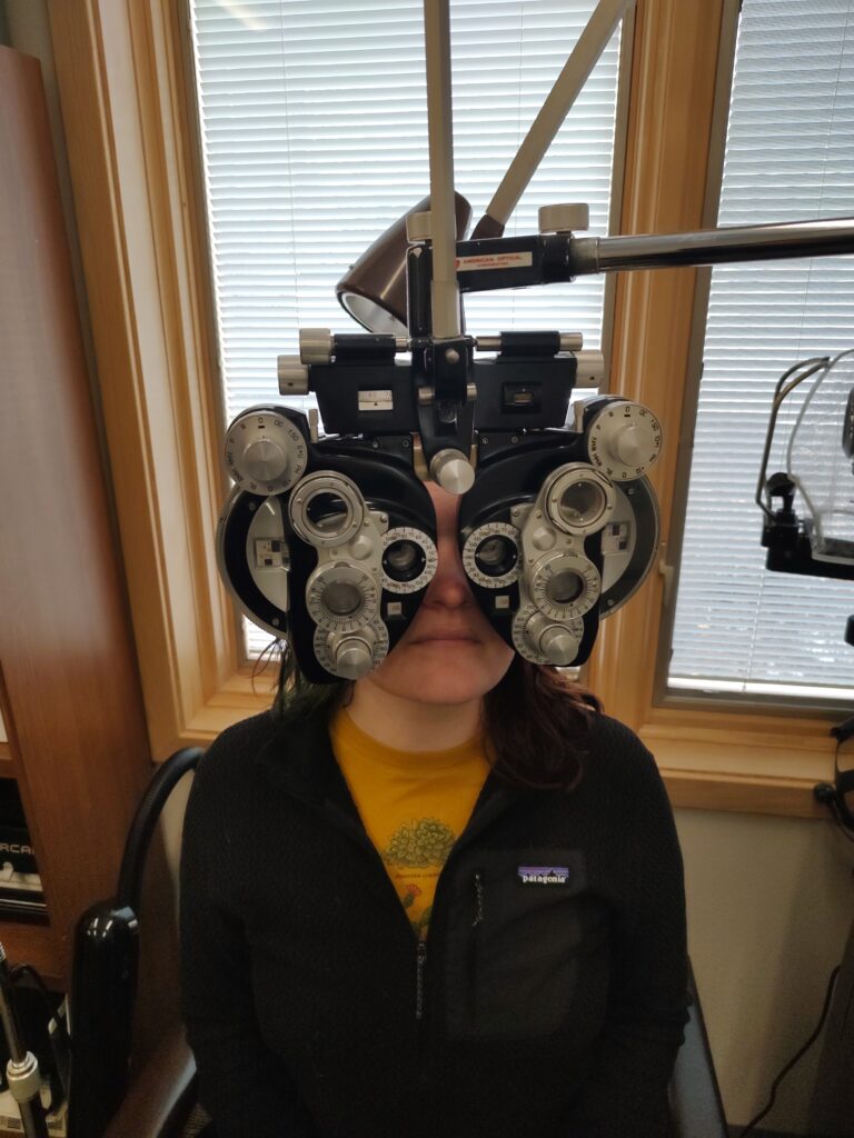 A patient looks through a phoropter during an eye examination. The person wears a yellow shirt and black Patagonia jacket while positioned behind the large optometry instrument, which features multiple rotating lens dials and adjustment knobs. The exam takes place in a bright office with windows covered by white blinds and wood trim visible in the background.