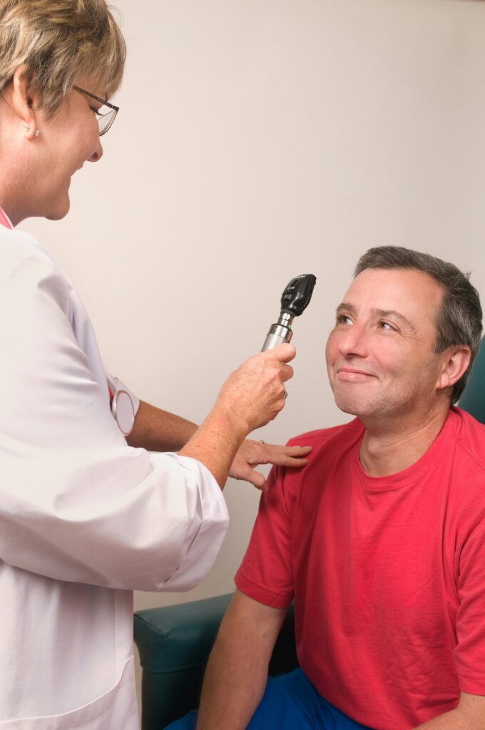 A healthcare provider in a white coat and glasses uses an ophthalmoscope to examine the eyes of a male patient wearing a red t-shirt. The patient is seated and looking upward while the doctor holds the diagnostic instrument near his face. The examination takes place against a neutral beige wall in a clinical setting.