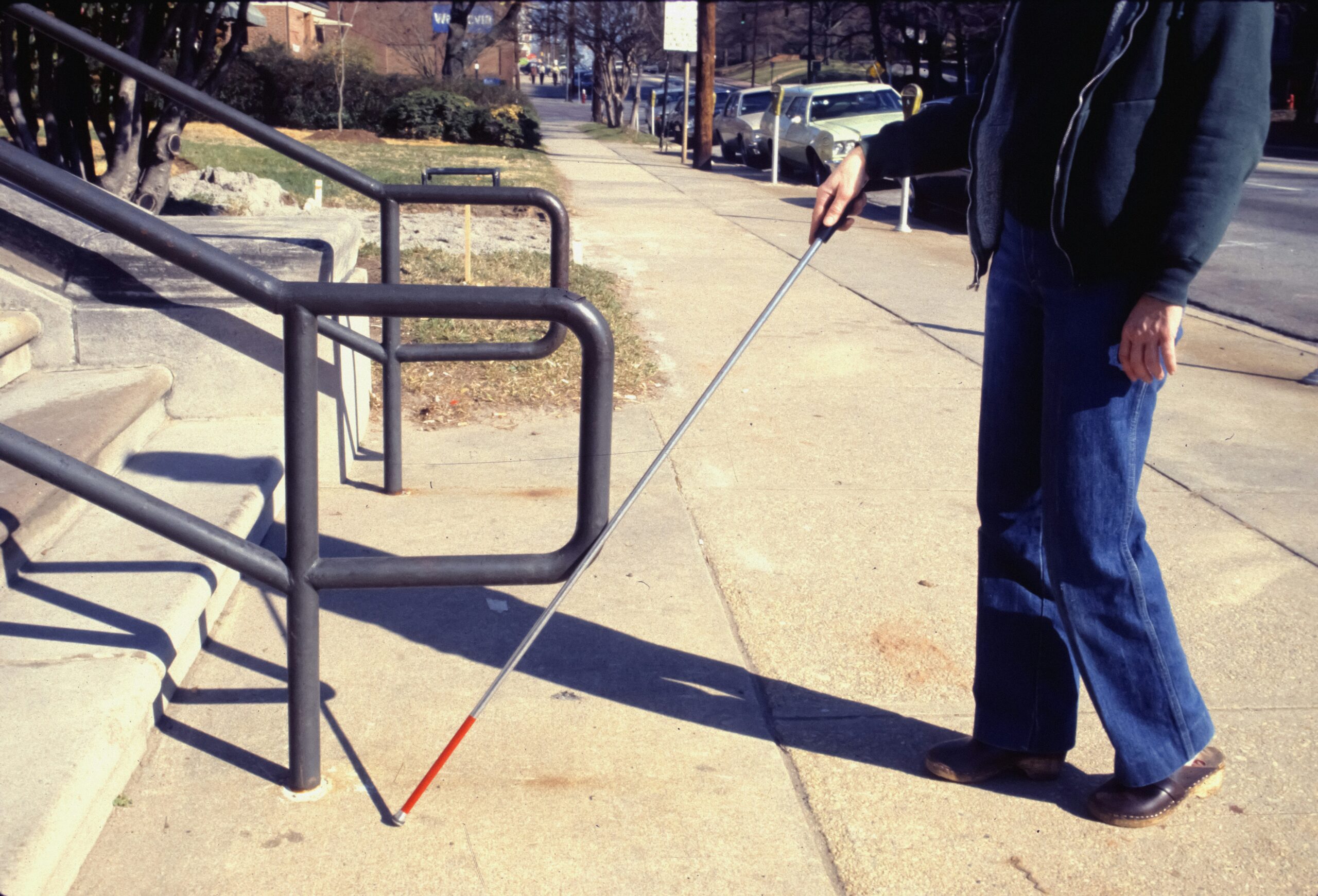 A person with a white cane with a red tip walks along a concrete sidewalk past metal handrails near steps. The person wears jeans and a dark jacket. In the background, a residential street is visible with parked cars, bare trees, grass, and buildings. The white cane is used for navigation and mobility assistance.