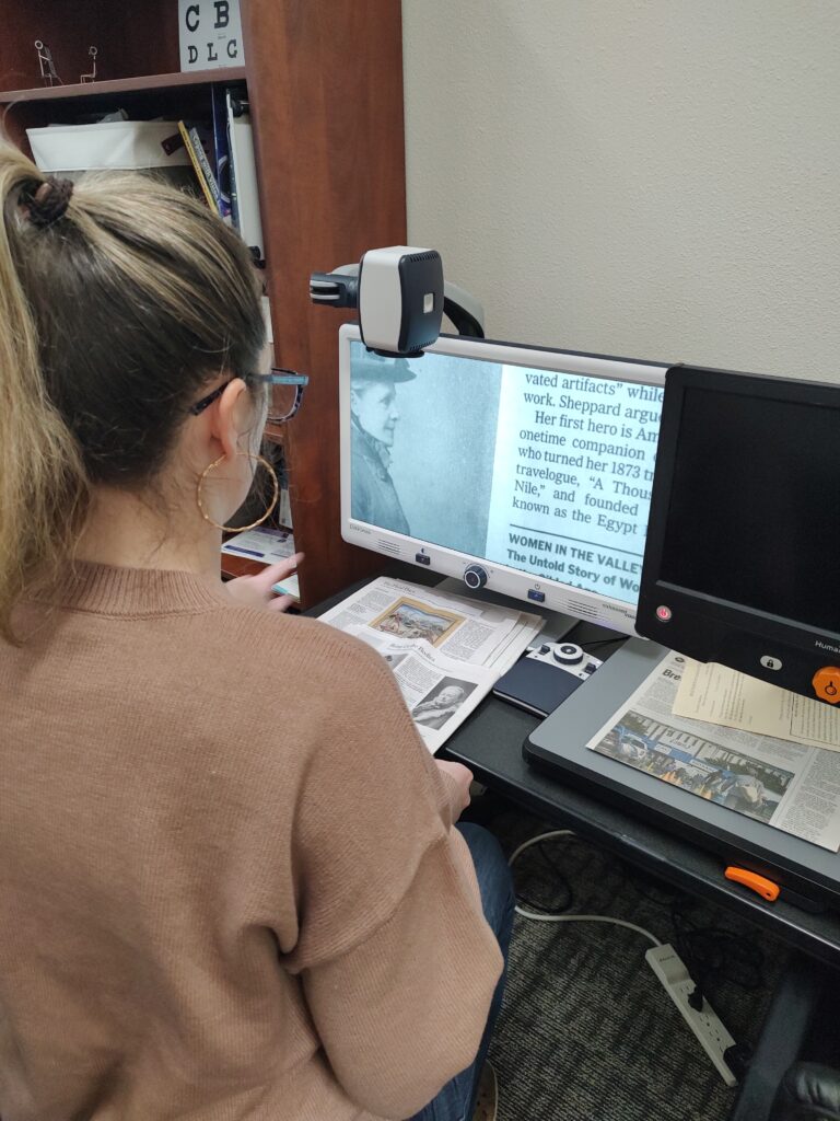 A woman wearing glasses and a tan sweater uses a video magnifier at a desk to read printed materials. The assistive technology device displays magnified text on a monitor showing content about women in history. Physical documents and newspapers are positioned on the reading platform beneath the camera. A bookshelf is visible in the background.
