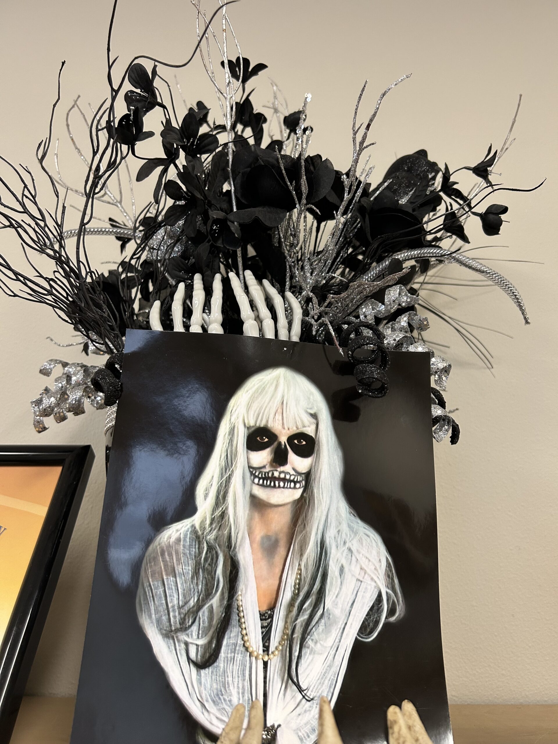Portrait photograph displayed on a wall showing a person with long white hair wearing dramatic skeleton face paint makeup. The portrait is topped with an elaborate Halloween decoration featuring black roses, white skeletal hands, twisted black branches, silver glittered twigs, and decorative elements creating a dramatic gothic centerpiece. The overall display combines photography and three-dimensional Halloween decor in black, white, and silver tones against a beige wall
