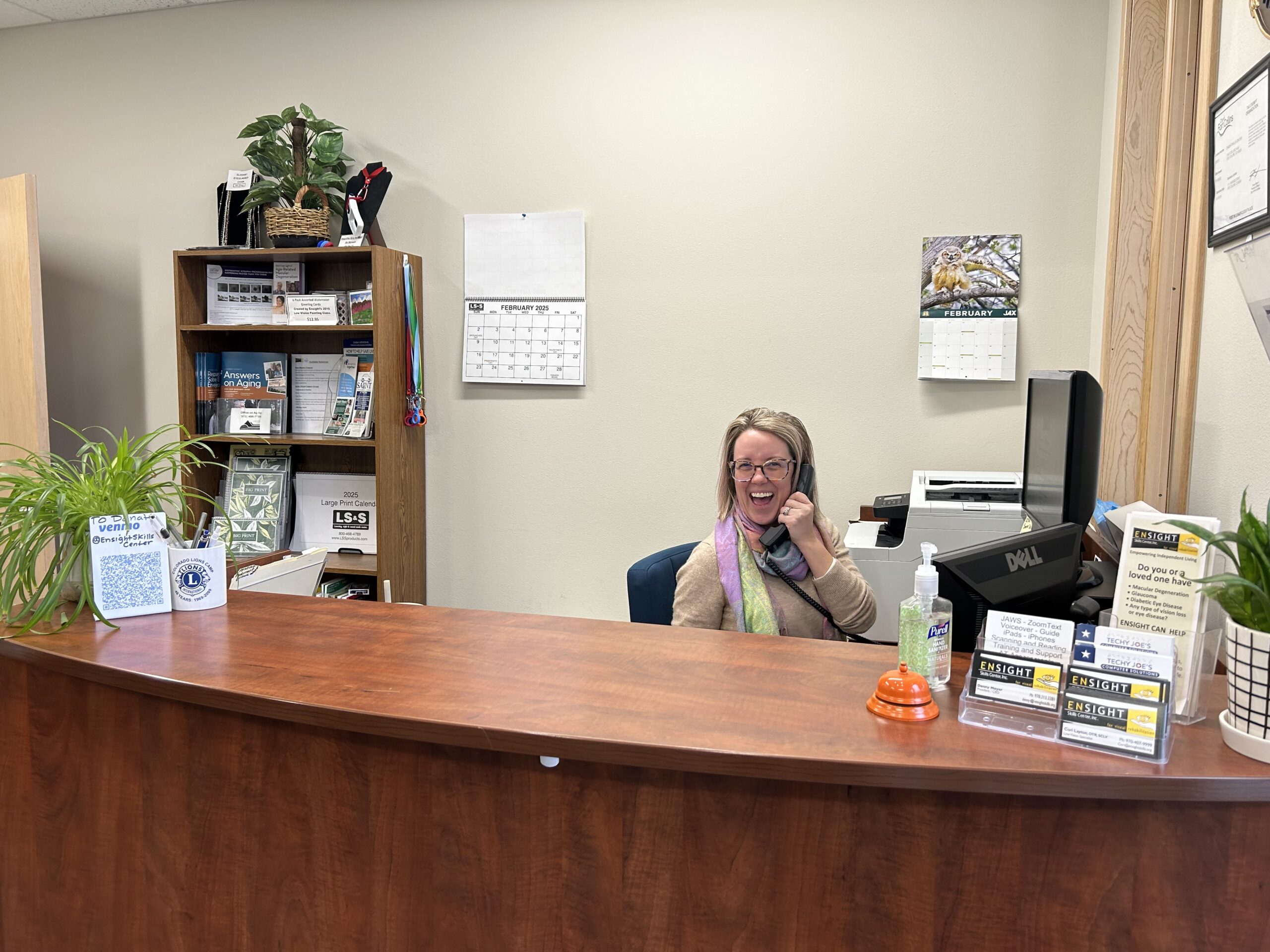 A smiling receptionist with glasses and shoulder-length blonde hair talks on a desk phone at a reception counter. She wears a tan cardigan and colorful scarf. The counter displays a hand sanitizer dispenser, business cards in an acrylic holder, and promotional materials. Behind her is a wooden shelving unit with office supplies, plants, wall calendars showing February, a Dell computer monitor, and cream-colored walls.