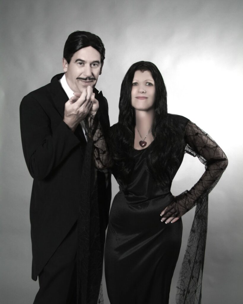 Black and white photo of a couple in formal Halloween attire. A man in a dark suit with a patterned vest poses with his hand near his chin, styled with slicked black hair and a mustache. Beside him, a woman in an elegant black lace dress with long sleeves and a heart-shaped necklace stands with one hand on her hip. Both are dressed in a classic Gothic or Addams Family-inspired style against a neutral backdrop.