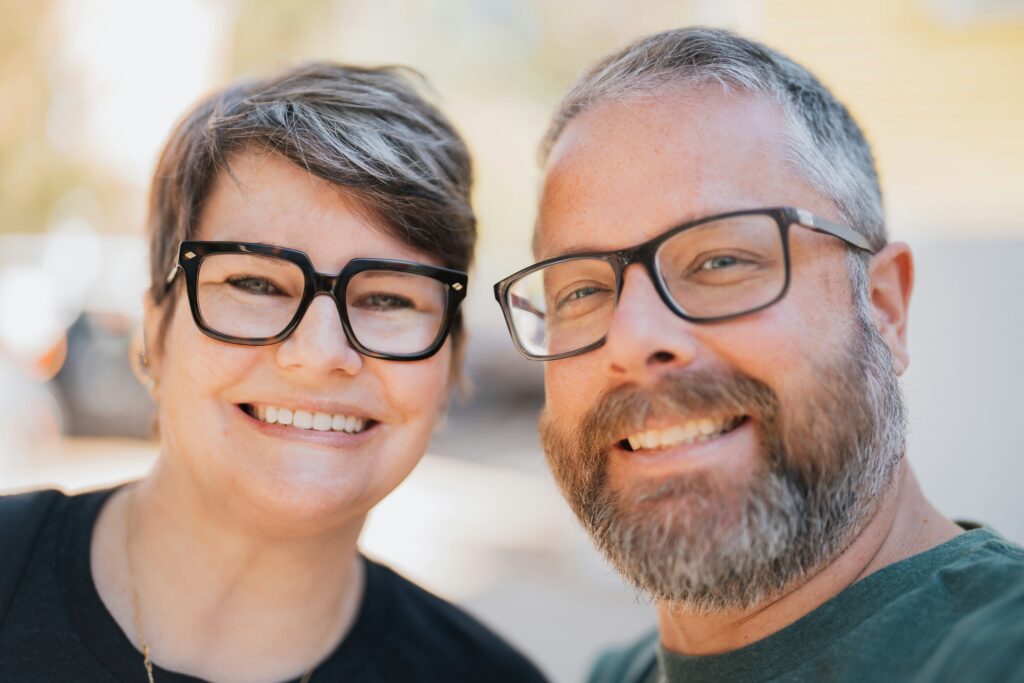 Two smiling adults wearing glasses pose together outdoors, with a soft, blurred background.