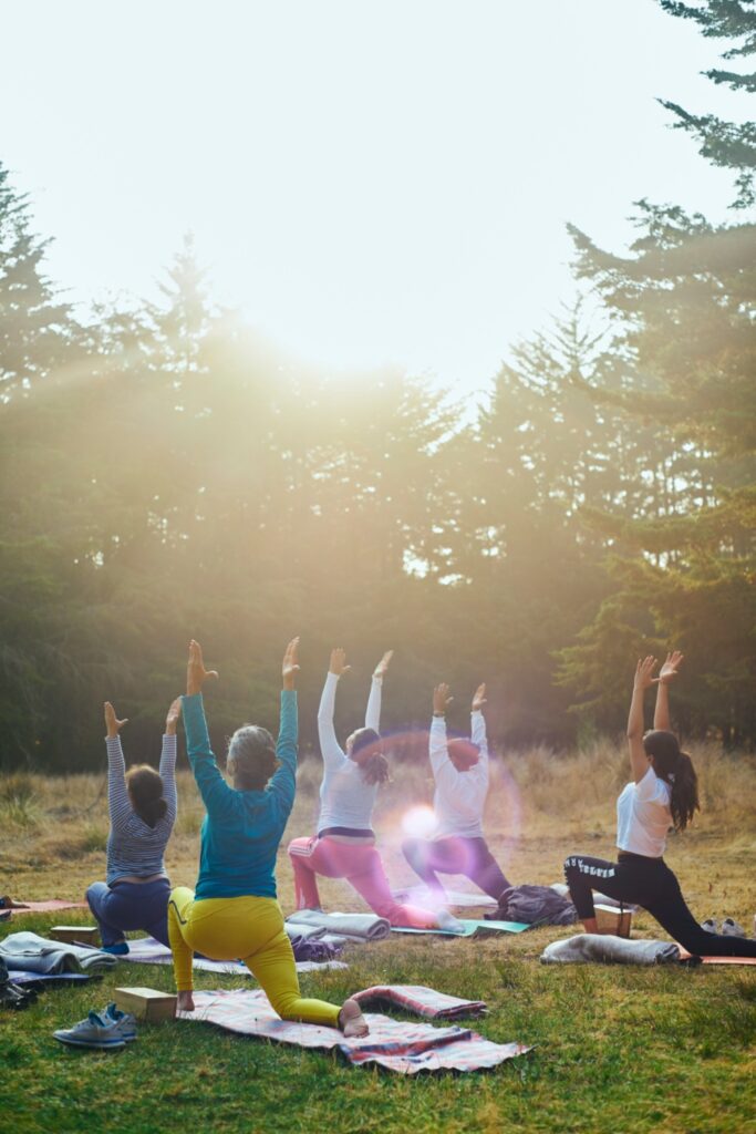 Several people participate in an outdoor yoga class on a grassy field, each on their own mat or blanket. They are in a low lunge pose — one knee on the ground, the other foot forward, with both arms extended upward. The group faces away from the camera toward a bright, hazy sun filtering through tall conifer trees. Participants wear colorful athletic clothing in teal, yellow, white, and striped patterns. Yoga blocks, shoes, and extra layers are visible at the edges of the mats.
