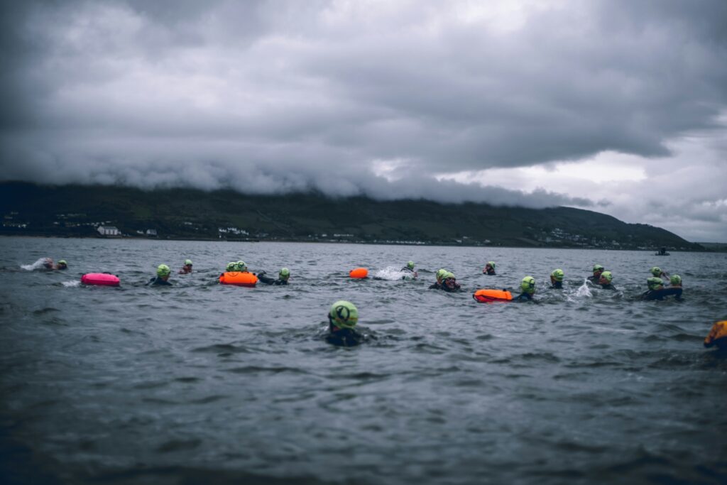A group of open-water swimmers in dark wetsuits and neon yellow-green swim caps make their way across a grey, choppy bay on a cloudy day. Many swimmers tow brightly colored safety buoys in orange and pink. The water is rough and the sky is heavy with dark clouds that partially obscure a green hillside and small coastal town in the background. The scene has a dramatic, moody atmosphere typical of cold-water open-water swimming events.