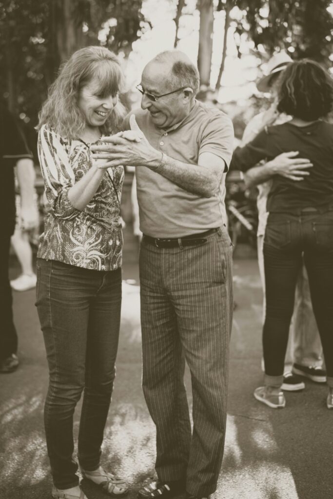 A sepia-toned photo of a man and woman dancing together outdoors on a paved surface, both laughing and smiling warmly. The woman has wavy hair and wears a patterned blouse and jeans; the man wears glasses, a polo shirt, and striped slacks. They hold hands between them in a partnered dance stance. In the background, another couple can be seen embraced in a similar dance. Trees with dappled light are visible in the soft-focus background.