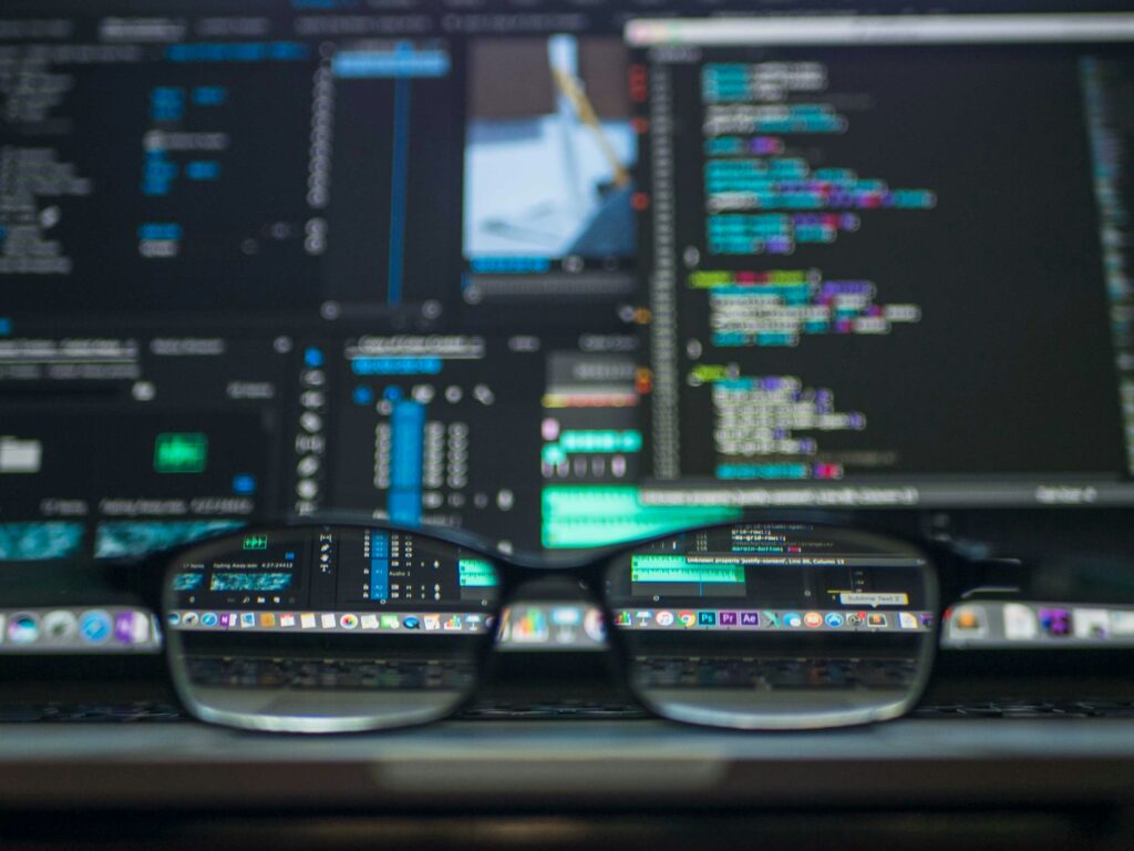 A pair of black-rimmed glasses resting in front of a computer monitor displaying colorful code and editing software.