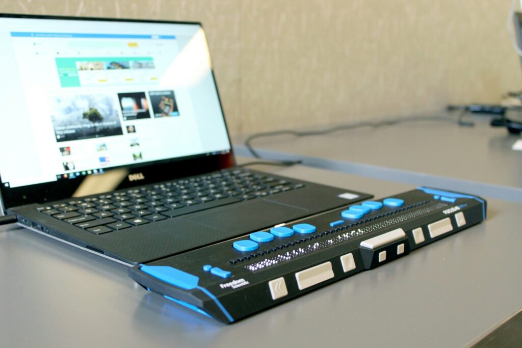 A Freedom Scientific refreshable braille display rests on a gray desk in front of an open Dell laptop. The device is black with bright blue and white control buttons and a row of tactile braille cells running across its length. The laptop screen behind it shows a web browser open to a news or media website. A cable and additional peripheral devices are partially visible in the background.