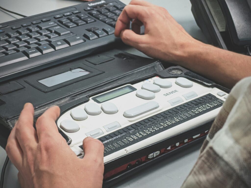 Close-up of a person's hands touching a refreshable braille display labeled 'SENSE,' which features a row of tactile braille cells, several rounded control buttons, and a small LCD screen. A standard black Microsoft keyboard sits on the desk just above the device. A monitor is partially visible in the upper right corner.