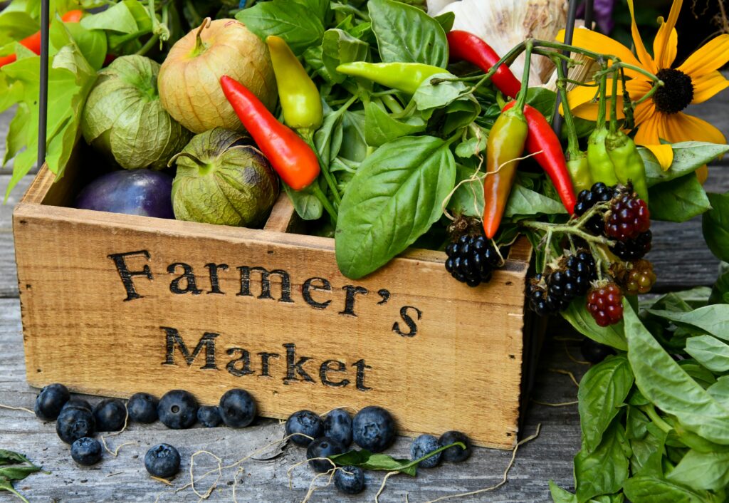Alt Text: A wooden crate labeled "Farmer's Market" overflowing with fresh produce, including tomatillos, chili peppers, basil, blackberries, and a purple plum, surrounded by scattered blueberries on a rustic wood surface.<