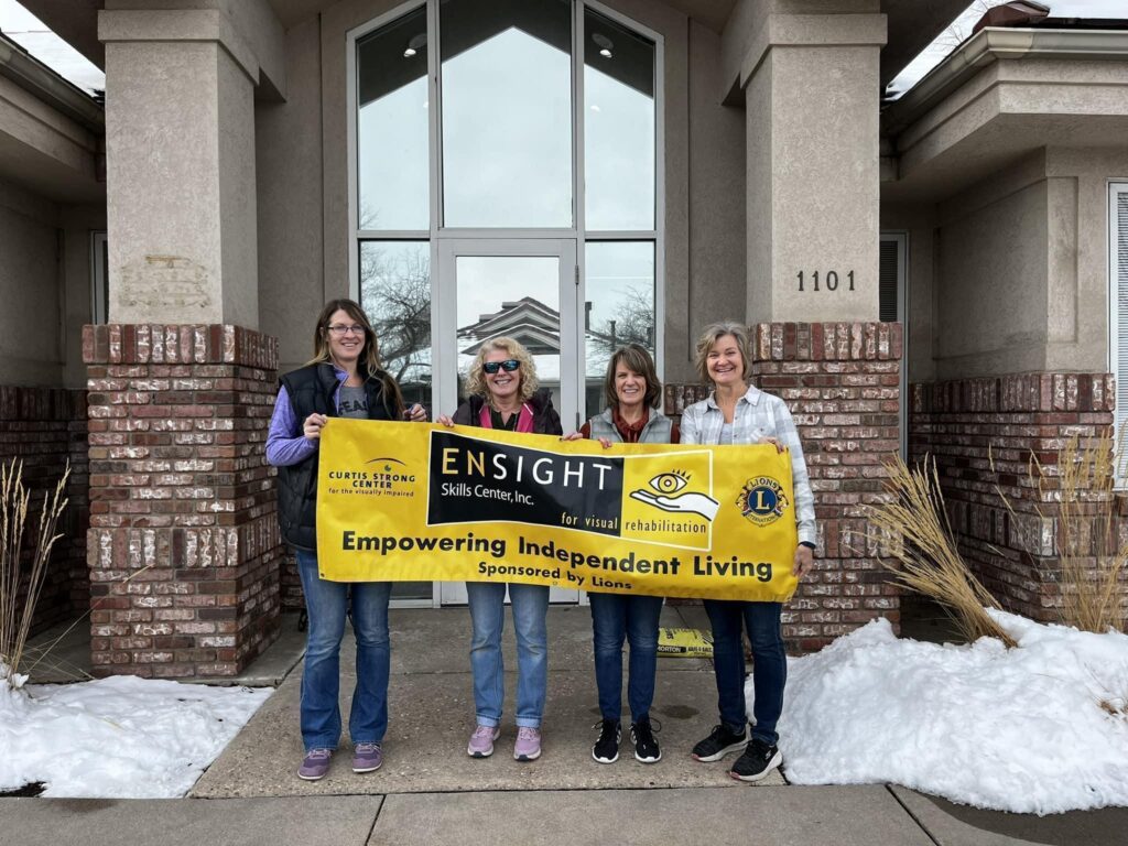 >ALT TEXT< The image shows four women standing outside a building in front of a doorway, holding a yellow banner with the text "ENSIGHT Skills Center, Inc. Empowering Independent Living" and logos for "Curtis Strong Center for the Visually Impaired" and "Lions International." The group is standing on a concrete path with patches of snow on the ground.
>ALT TEXT< The image shows four women standing outside a building in front of a doorway, holding a yellow banner with the text "ENSIGHT Skills Center, Inc. Empowering Independent Living" and logos for "Curtis Strong Center for the Visually Impaired" and "Lions International." The group is standing on a concrete path with patches of snow on the ground.