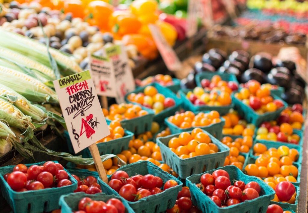 >Alt Text: Rows of red and yellow cherry tomatoes in green cartons at a farmers market stand, with a hand-lettered "Organic Fresh Cherry Tomatoes $4/box" sign, fresh corn, and other colorful produce in the background.<