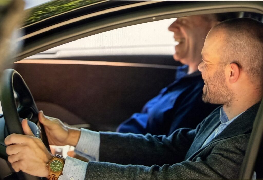 A man drives a car while smiling and talking with an older man seated in the back seat, seen through the open front window with one hand on the steering wheel.