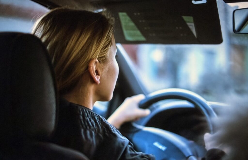 A woman with shoulder-length blonde hair drives a car, seen from behind the driver's seat, one hand on the steering wheel as she looks out at the road ahead.