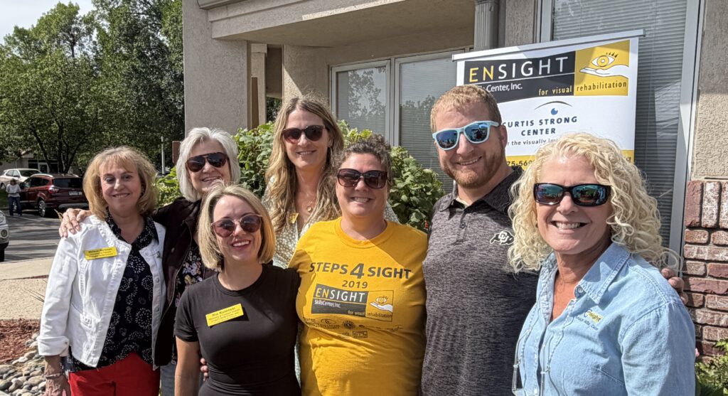 Seven Ensight Skills Center staff and supporters stand shoulder to shoulder outdoors on a sunny day, smiling at the camera, most wearing sunglasses. Behind them hang an Ensight Skills Center banner reading “Curtis Strong Center for the visually impaired” and a leafy green shrub. Two of the women wear yellow name tags reading Melissa Workman and Ria Kulenski. One wears a yellow “Steps 4 Sight 2019” Ensight T-shirt, one a light denim shirt with the Ensight logo, and a man in teal sunglasses wears a gray University of Colorado polo.