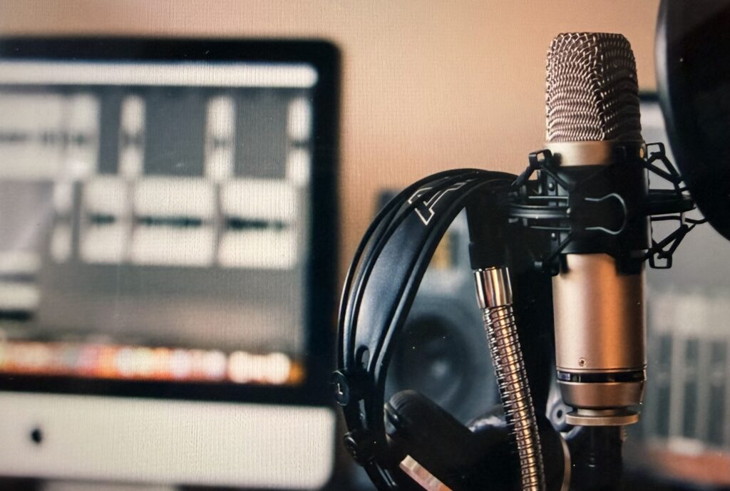 A studio microphone in a shock mount with a pair of black headphones hanging from it, in front of a blurred computer screen showing audio editing software.