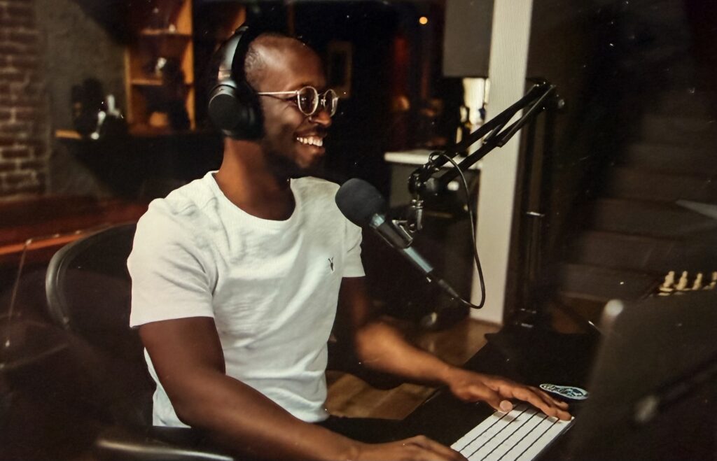 A smiling man wearing over-ear headphones and glasses sits at a desk in front of a microphone on a boom arm, with one hand on a white keyboard, as if recording a podcast.