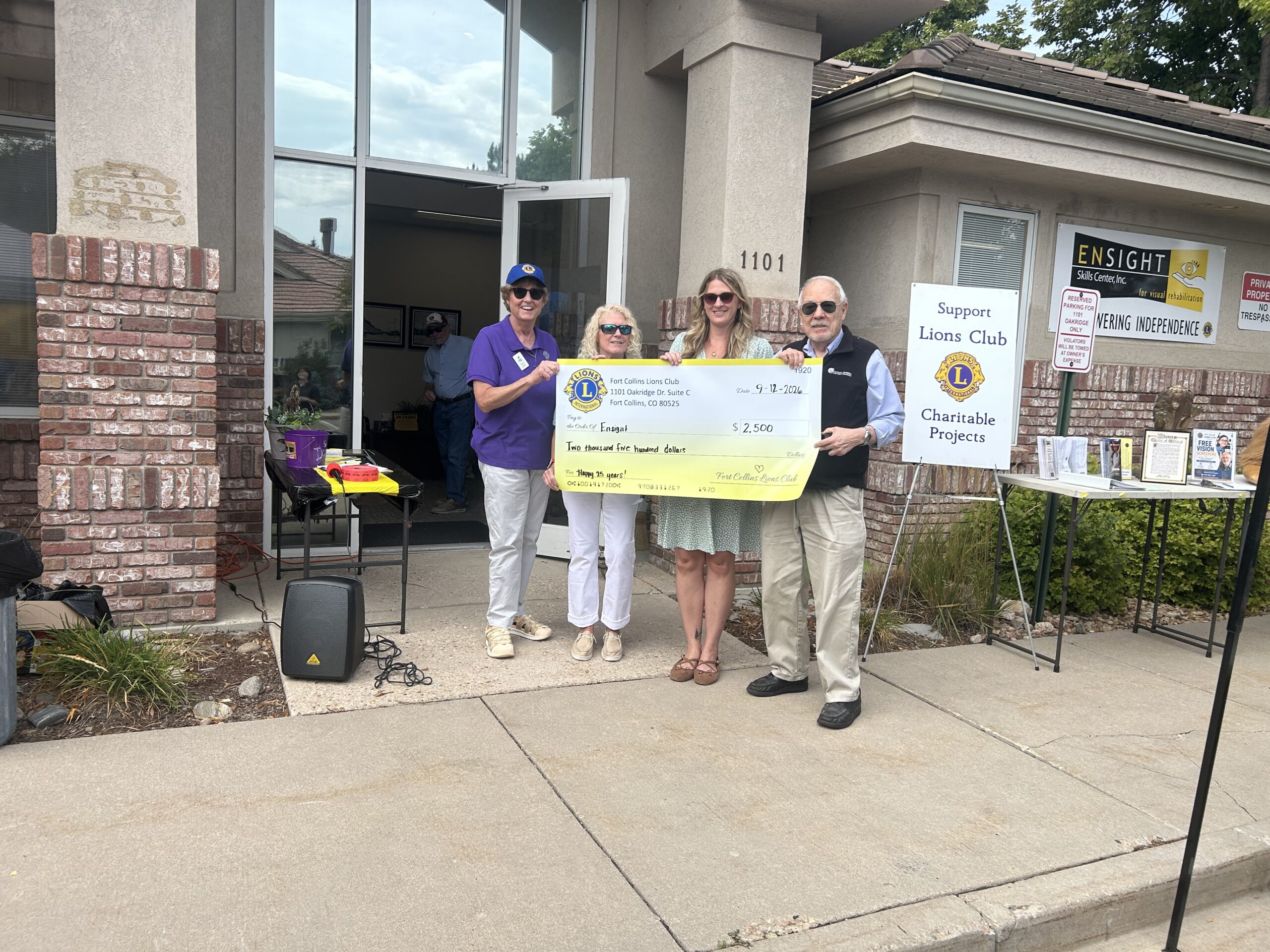 Four smiling people stand outside the entrance of Ensight Skills Center holding a large yellow presentation check from the Fort Collins Lions Club. The check is dated 9-12-2026, is made out to Ensight for $2,500 (two thousand five hundred dollars), and has “Happy 25 years!” written on the memo line. Two women on the left, one in a purple Lions Club polo and blue cap, stand beside a woman in white pants, a woman in a green patterned dress, and a man in a black vest and khakis. Behind them are the Ensight Skills Center sign, a “Support Lions Club Charitable Projects” sign, and a table with informational materials.